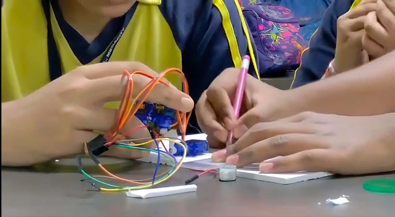 Hands wiring the servos of the mini humanoid robot on a workbench, with colourful jumper wires and a pencil marking the base board