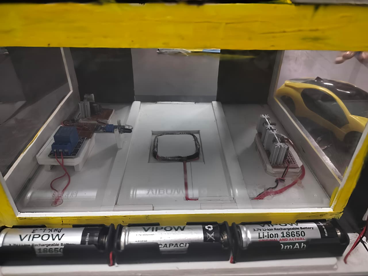 The wireless charging EV model: a charging pad with its coil in the middle of a road track, relay and regulator boards on either side, a yellow model car at the end and a row of 18650 lithium cells along the front